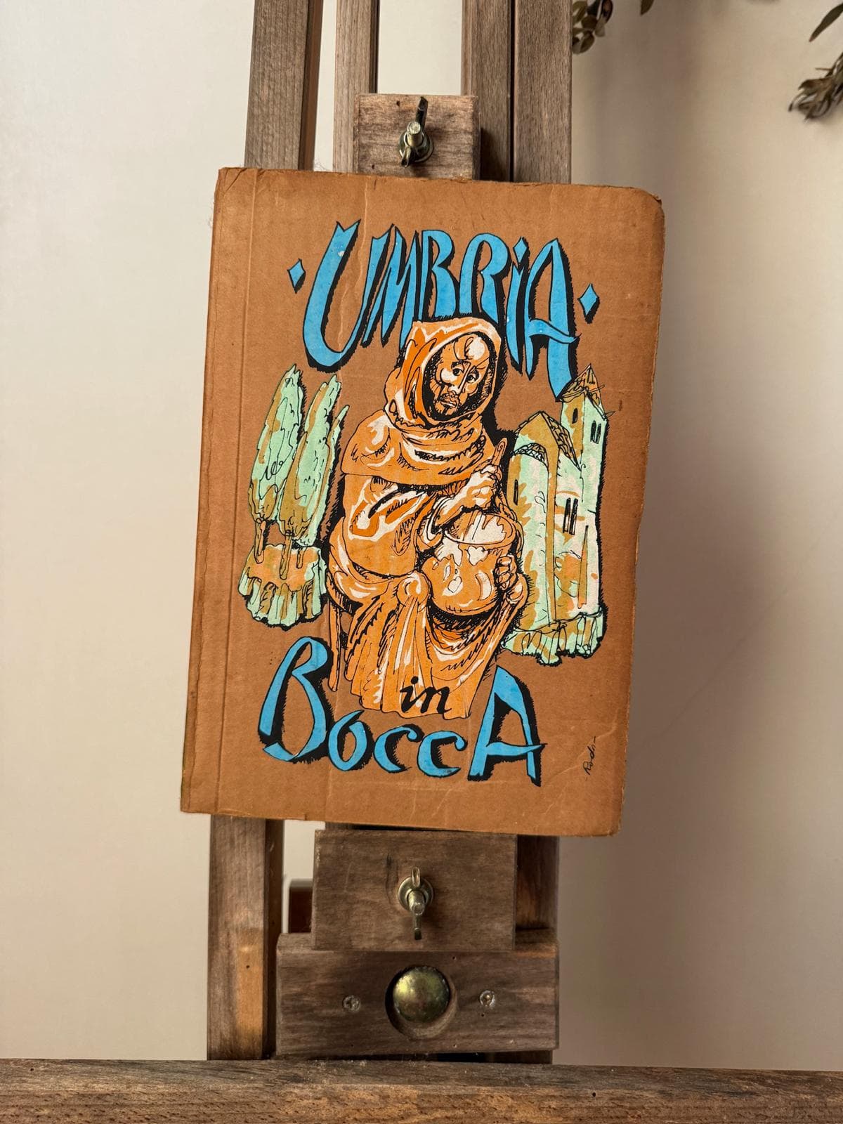 A picture of Umbria in Bocca a rare and collectible Vintage Cook Book for sale from the Jolene Bakery. The picture shows the cover of the book, a hand painted illustration depicting a man in a hooded brown cloak mixing food, some trees and a stone building