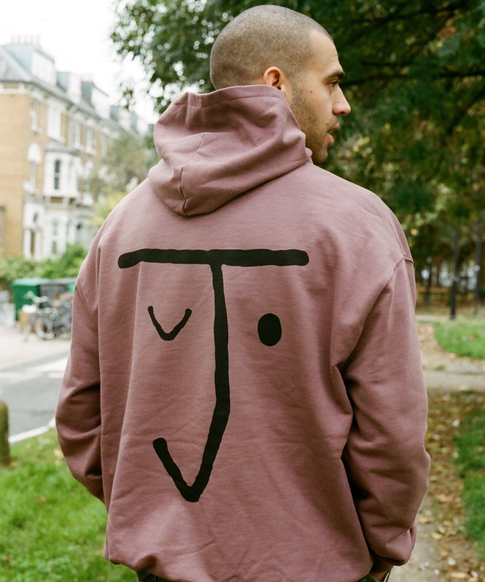 A male model standing outside with his back to the camera wearing the Jolene bakery J face hoodie in coffee