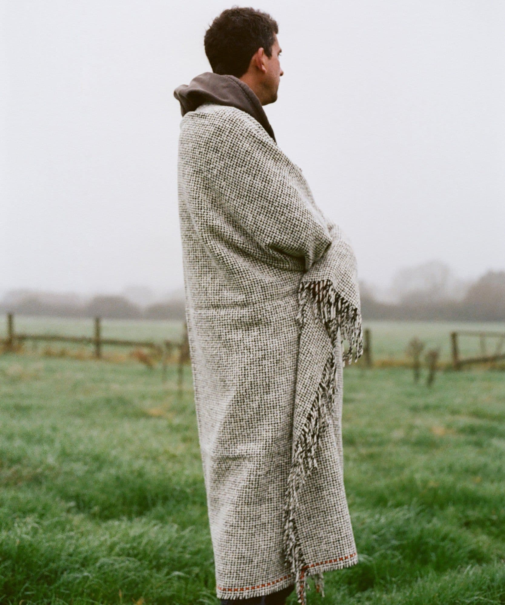 A model wrapped up in the giant Jolene x Mourne Textiles blanket standing in a green field in the winter, it is quite a grey and misty scene