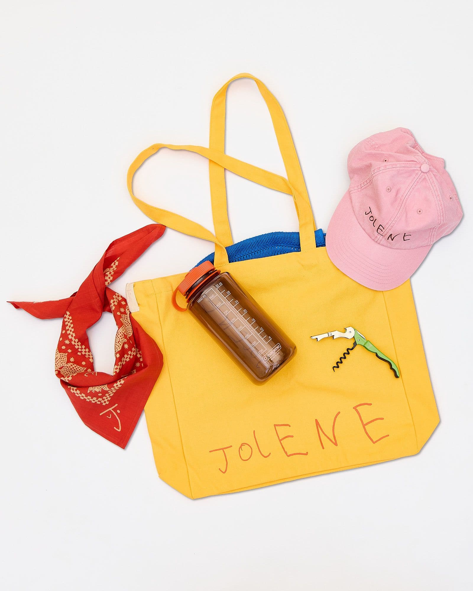 A picture of the Jolene bakery refillable water bottle with the Jolene yellow tote bag, Jolene green bottle opener, Jolene pink baseball cap and Jolene red scarf