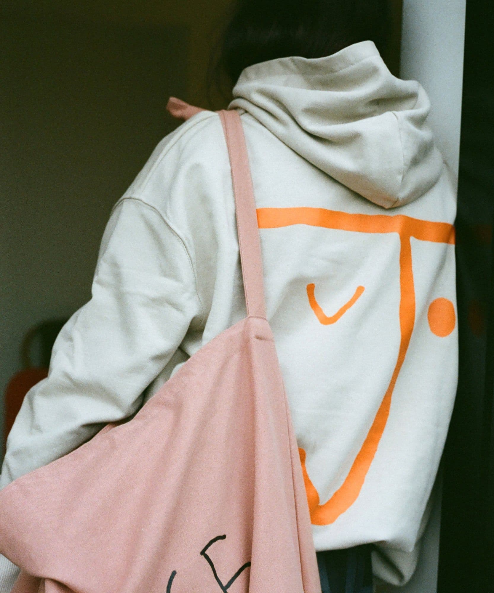 A model wearing the J face hoodie in stone with a bright orange J face logo on the back. On her shoulder is the Jolene bakery big bag