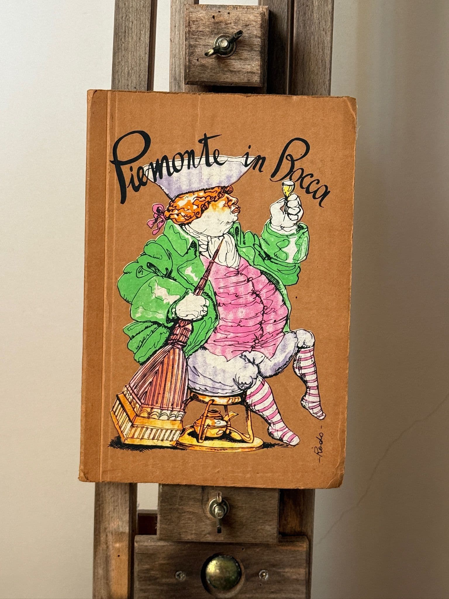 A picture of Piemonte in Bocca a rare and collectible Vintage Cook Book for sale from the Jolene Bakery. The picture shows the cover of the book, an hand painted illustration depicting a large man in a pink waistcoat, green coat, red and white stockings drinking a glass of wine and looking rather merry